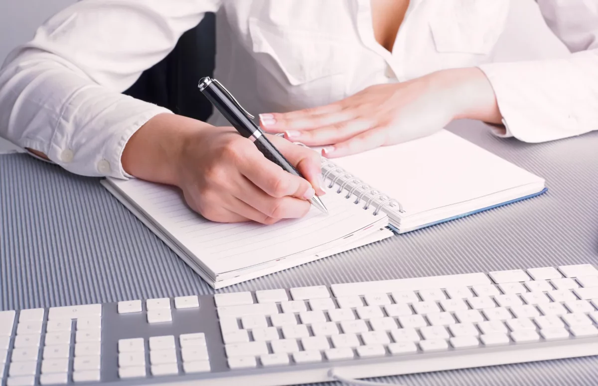 femme qui écrit sur un cahier assise à un bureau sur lequel on voit un clavier d'ordinateur pour symboliser les services de rédaction web que je propose